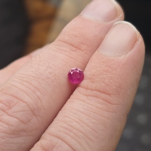 Puede incluir: Una gema de rubí facetada y redonda. La gema es de color rojo vibrante y se sostiene entre dos dedos. La gema tiene múltiples facetas que reflejan la luz, dándole un aspecto brillante. La gema es pequeña, probablemente menos de 1 quilate.