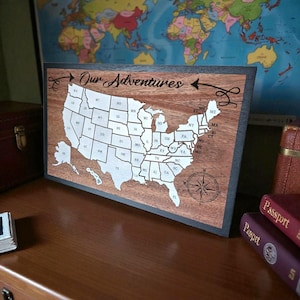May include: Wooden map of the United States with state outlines in white. The map is framed in black and features the text "Our Adventures" and a compass rose. A world map is in the background.