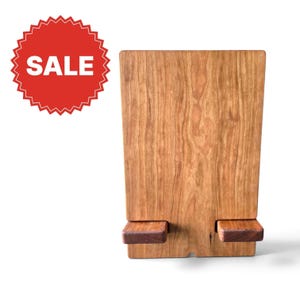 May include: A rectangular wooden stand featuring a smooth, light brown grain pattern with two small, horizontal wooden supports attached to the lower front section. A red circular badge with a serrated edge displays the white text SALE.