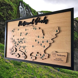 May include: Wooden map of the United States with a black frame, featuring a detailed outline of each state. The map includes small tree icons marking national parks and the text "National Parks - Explore the Outdoors". A smaller plaque reads "Welcome to our Adventure".