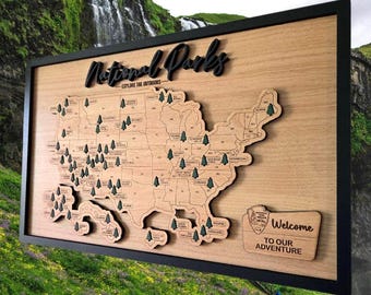 May include: Wooden map of the United States with a black frame, featuring a detailed outline of each state. The map includes small tree icons marking national parks and the text "National Parks - Explore the Outdoors". A smaller plaque reads "Welcome to our Adventure".