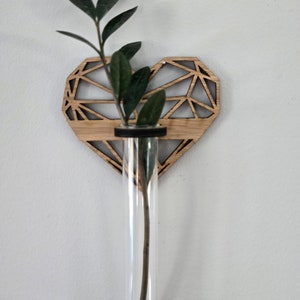 May include: A wooden heart-shaped vase with a geometric design. A clear glass test tube is inserted into the vase, holding a single green stem with leaves.