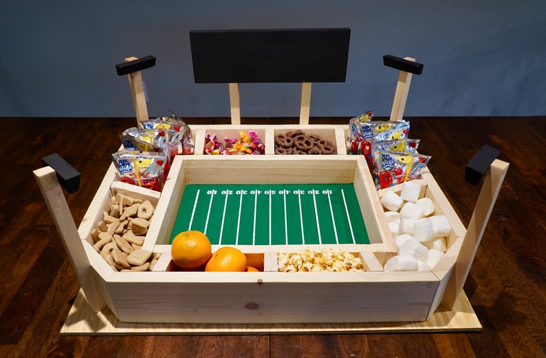 May include: A wooden football stadium shaped snack serving tray with a green field and white yard lines. The tray has six compartments filled with various snacks including pretzels, marshmallows, orange slices, popcorn, and bags of chips.