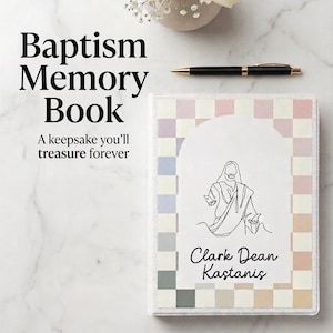 May include: A Baptism Memory Book with a pastel-coloured chequered pattern. The cover displays a line drawing of a religious figure and the name "Clark Dean Kastanis". The book is on a marble surface, with a pen and flowers nearby.