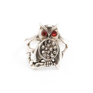 Peut inclure: Bague en argent représentant un hibou avec des yeux en pierres rouges. Le hibou présente un design détaillé avec un corps texturé et un ventre rond. La bague est posée sur un fond blanc.