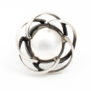 May include: A silver ring with a large, white pearl at its centre. The pearl is held within a decorative, openwork design of curved, interlocking metal bands. The ring's design is symmetrical and elegant, suitable for formal or everyday wear.