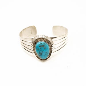 May include: Silver cuff bracelet featuring a large, oval turquoise stone. The turquoise is set in a silver bezel and surrounded by a twisted rope design. The bracelet has a simple, yet elegant design.