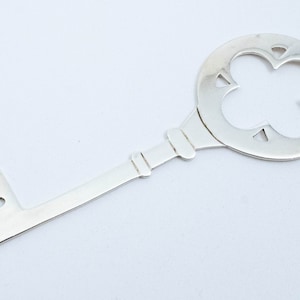 May include: A silver key pendant with a decorative, openwork, four-leaf clover design. The key has a loop at the top for attaching to a chain.