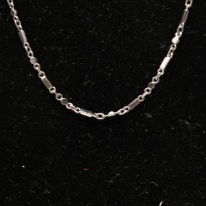 May include: A delicate silver necklace with a repeating pattern of small rectangular and circular links. The chain is displayed against a dark background, highlighting its reflective surface. This piece is suitable for everyday wear.