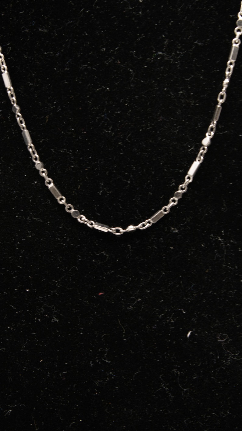 May include: A delicate silver necklace with a repeating pattern of small rectangular and circular links. The chain is displayed against a dark background, highlighting its reflective surface. This piece is suitable for everyday wear.