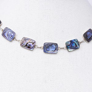 Peut inclure: Un collier composé de sept maillons rectangulaires en nacre d'ormeau reliés par des anneaux en métal argenté. Chaque maillon présente un motif irisé unique avec des nuances tourbillonnantes de bleu, violet, vert et gris, disposés en ligne.