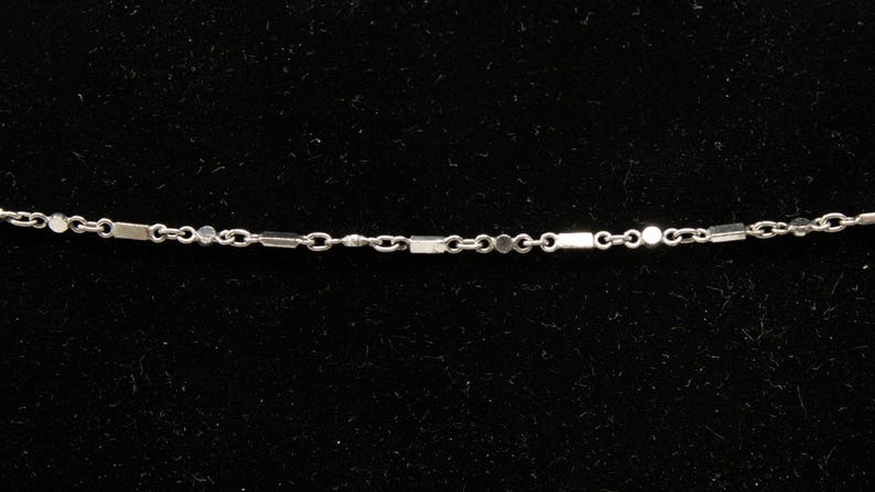 May include: Delicate silver necklace with a chain featuring alternating rectangular and circular links. The necklace is displayed against a black background, highlighting its minimalist design. A fine piece of jewelry.