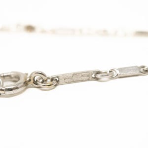 May include: Close-up of a silver necklace clasp and chain. The chain features small rectangular and round links. The clasp is a circular spring ring type. The necklace is set against a white background.