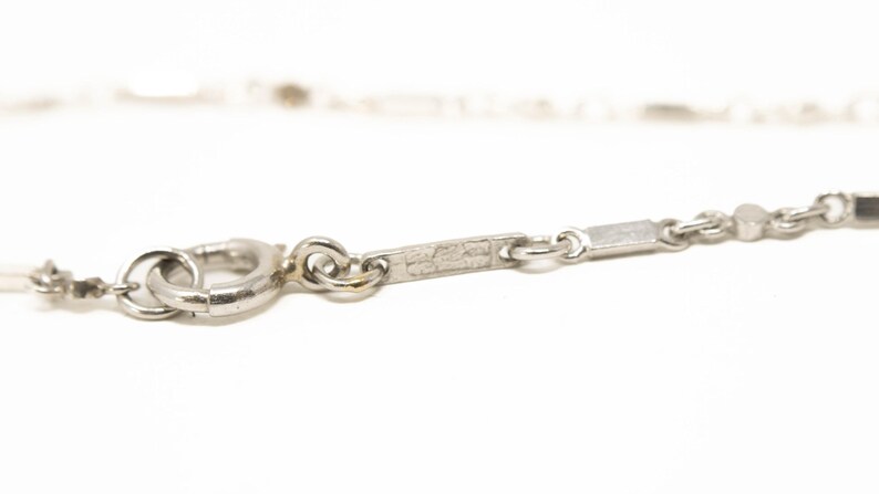 May include: Close-up of a silver necklace clasp and chain. The chain features small rectangular and round links. The clasp is a circular spring ring type. The necklace is set against a white background.