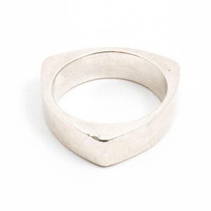 Könnte beinhalten: Ein silberfarbener Ring mit dreieckiger Form. Der Ring hat eine flache Oberseite und Seiten mit abgerundeten Ecken. Der Ring besteht aus massivem Metall und hat eine glatte, matte Oberfläche.