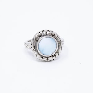 May include: A silver ring featuring a pale blue, oval-shaped central stone. The ring is decorated with an intricate, openwork pattern around the stone and on the band. Set against a white backdrop.