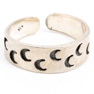 May include: A silver adjustable ring with a crescent moon pattern. The ring has a series of crescent moon cutouts around the band. The ring is open at the top for size adjustment. The metal has a slightly aged appearance.