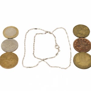 May include: A silver necklace with a delicate chain design, positioned between three stacks of coins. The coins vary in color, including gold, silver, and bronze, and are arranged in vertical rows.