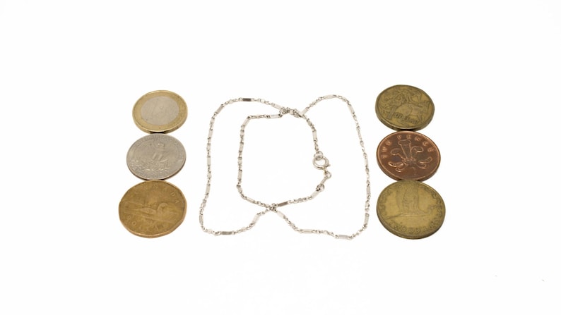 May include: A silver necklace with a delicate chain design, positioned between three stacks of coins. The coins vary in color, including gold, silver, and bronze, and are arranged in vertical rows.