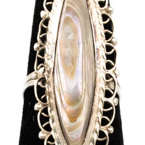 Op de afbeelding: Zilveren ring met een ovale, iriserende abalone schelp middensteen. De schelp toont een mix van crème, bruin en parelmoer kleuren. De ring heeft een decoratieve, opengewerkte rand en een eenvoudige band.