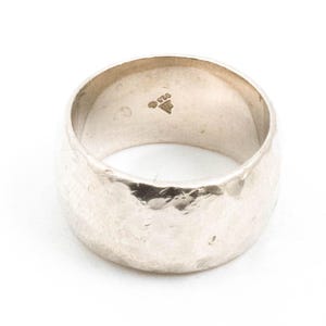 Può includere: Un anello d'argento con una texture martellata. L'interno della fascia è timbrato con il numero "925" e un segno che assomiglia a un triangolo.