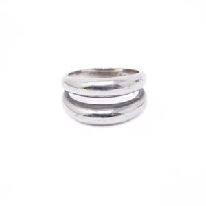 May include: A polished silver-toned ring featuring a double-band design. The ring consists of two smooth, rounded, parallel bands connected at the sides, creating a distinct open space between the upper and lower sections of the metal band.