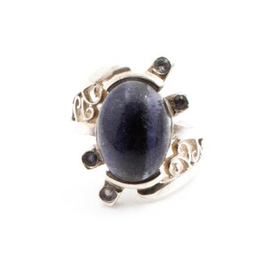Può includere: Un anello d'argento con una grande pietra preziosa ovale blu scuro. L'anello presenta un elaborato motivo a spirale sulla fascia e piccole pietre scure attorno alla pietra principale. L'anello è su sfondo bianco.