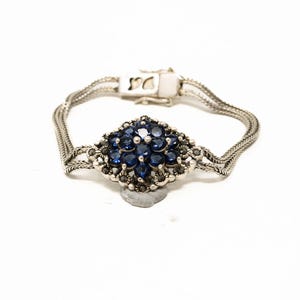 May include: Silver bracelet featuring a central cluster of deep blue gemstones surrounded by smaller, dark stones. The bracelet has a woven chain design and a secure clasp. A piece of jewelry.