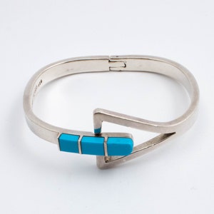 Può includere: Un bracciale argentato con cerniera e design geometrico. Il bracciale presenta tre pietre rettangolari color turchese. La chiusura è di design semplice. Un gioiello adatto a diverse occasioni.