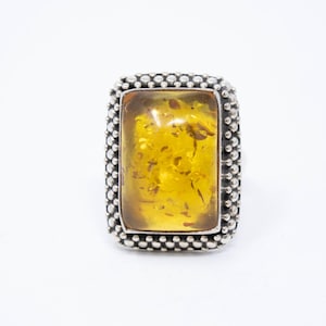 Könnte beinhalten: Rechteckiger Bernsteinring mit silberner Perlenbordüre. Der Bernsteinstein ist ein durchscheinendes Gelb mit sichtbaren Einschlüssen. Der Ring ist vor weißem Hintergrund platziert.
