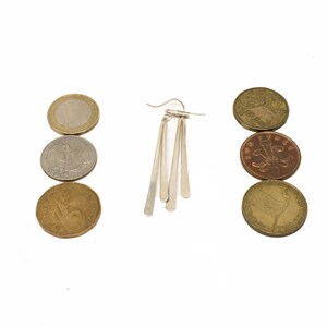Pu&ograve; includere: Un orecchino d'argento con tre pendenti allungati a forma di goccia &egrave; centrato su uno sfondo bianco. Sei monete di varie dimensioni e colori sono disposte in due colonne verticali, ai lati dell'orecchino.