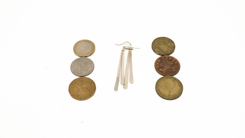 Pu&ograve; includere: Un orecchino d'argento con tre pendenti allungati a forma di goccia &egrave; centrato su uno sfondo bianco. Sei monete di varie dimensioni e colori sono disposte in due colonne verticali, ai lati dell'orecchino.