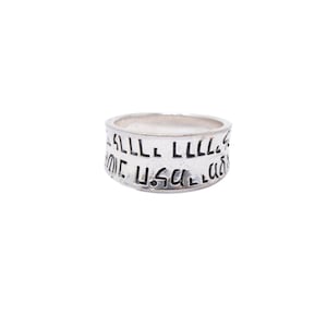 May include: A silver-toned band ring featuring a smooth, polished finish. The exterior surface of the ring is engraved with a series of black, stylized, hand-drawn characters and symbols that wrap around the entire circumference of the metal band.