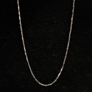 May include: A delicate silver necklace with a bar and chain design. The necklace is displayed against a black background, highlighting its simple elegance. The chain is approximately 18 inches long.