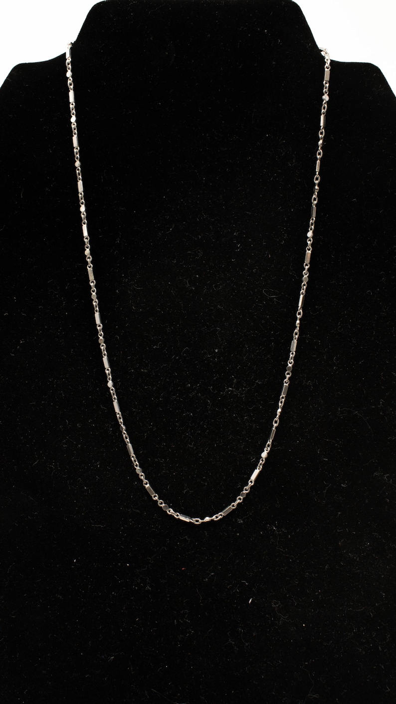 May include: A delicate silver necklace with a bar and chain design. The necklace is displayed against a black background, highlighting its simple elegance. The chain is approximately 18 inches long.