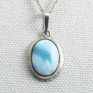May include: A silver pendant necklace featuring an oval-shaped, light blue gemstone. The gemstone is set in a silver bezel and hangs from a delicate silver chain. The pendant has a polished finish and is displayed against a neutral background.