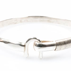 May include: A silver bangle bracelet with a hook clasp. The bracelet has a hammered texture and a coiled wire detail near the clasp. The jewellery is displayed against a white background.