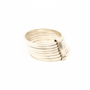 May include: A silver ring with a distinctive design. The ring comprises several stacked bands secured by a single, broader band. The ring is set against a white backdrop.
