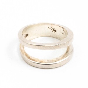 May include: A silver ring featuring a double-band design. The ring has a smooth, polished finish and a minimalist style. The two bands are parallel and connected, creating a unique and modern look. The ring is set against a white background.