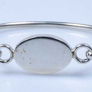 May include: A silver bangle bracelet with a smooth oval-shaped charm. The charm is blank and can be engraved.