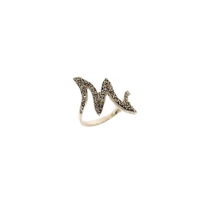 May include: A silver ring with a stylized letter "M" design. The letter is encrusted with small, dark-colored stones, creating a textured appearance. The ring has a polished, smooth band.