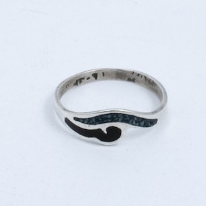Könnte beinhalten: Ein silberner Ring mit einem schwarzen und türkisfarbenen Einlegemuster. Der Ring ist ein schlichtes Band mit einem geschwungenen Design auf der Oberseite. Die Einlage ist eine wellenförmige Linie mit einer schwarzen Mitte und türkisfarbenen Rändern.