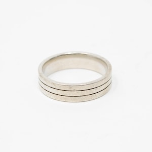 Peut inclure: Une bague argentée avec trois fines rainures parallèles autour du cercle. La bague est présentée sur un fond blanc uni, soulignant son design simple mais élégant. Ce bijou convient au port quotidien.