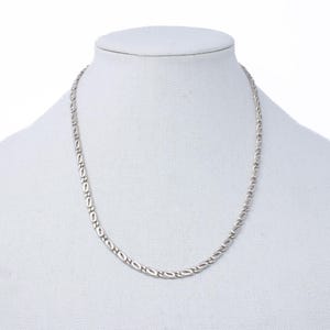 May include: A silver-colored necklace displayed on a white mannequin. The necklace features a repeating pattern of elongated oval links. The chain is delicate and appears to be made of metal. The background is plain white.
