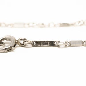 May include: Close-up of a silver necklace with a clasp and rectangular links. The necklace is on a white background. The clasp is a circular ring. The necklace has a small rectangular tag with markings.