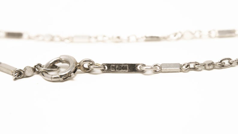 May include: Close-up of a silver necklace with a clasp and rectangular links. The necklace is on a white background. The clasp is a circular ring. The necklace has a small rectangular tag with markings.