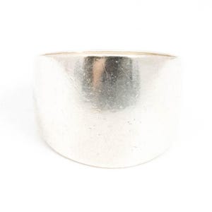 May include: A wide, silver ring with a smooth, polished surface. The ring has a cylindrical shape and a solid band. The metal reflects light, creating highlights and shadows. The ring is set against a plain white background.