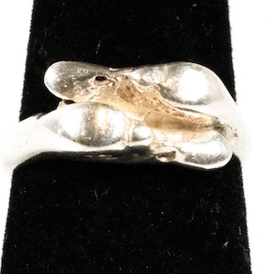 May include: A silver-colored ring with an abstract design. The ring features a series of rounded, organic shapes, possibly cast from a natural form. The ring is displayed against a black background, highlighting its metallic sheen.
