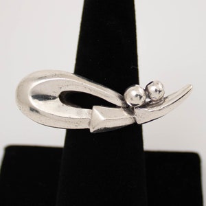 May include: A silver ring with a unique abstract design featuring a curved shape and two small spheres. The ring is displayed on a black stand.