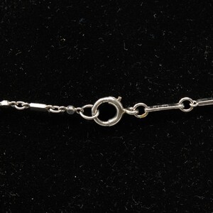 May include: Close-up of a silver necklace clasp and chain against a black background. The chain features a delicate design with small, rectangular links and circular connectors. The clasp is a simple, secure design.